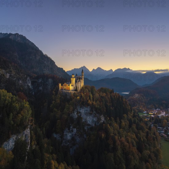 Illuminated Neuschwanstein Castle sits majestically on a wooded hill surrounded by mountains at dusk, Schwangau near Füssen, Ostallgäu, Allgäu, Bavaria, Germany