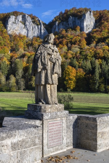 Statue, saint statue of Saint Nepomuk, patron saint of bridges, bridge saint on the historic arched bridge, Hausener Brücke behind the distinctive Hausener Zinnen, climbing rocks, Jurassic limestone rocks, Weissjura, Jurassic rocks, limestone rocks, surrounded by autumnal vegetation, Hausen im Tal, Upper Danube Valley, Baden-Württemberg, Germany
