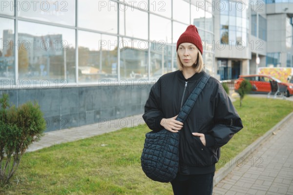 A woman wearing a black jacket and red beanie walks along a modern glass building. She holds a large bag and seems to enjoy a day in the city, surrounded by greenery and urban scenery
