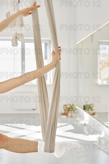 In a bright room with large windows, a person is using a hammock to perform fitness stretching. This exercise helps improve flexibility and strength through controlled movements using the fabric