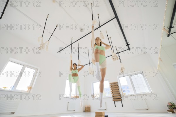 Two individuals engage in a fitness stretching routine using a hammock in a bright, spacious studio. The atmosphere is energetic as they focus on flexibility and strength training