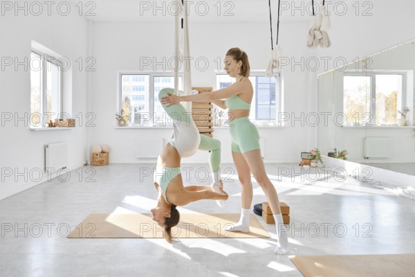 Two women engage in fitness stretching using a hammock in a spacious, well-lit studio. The environment is airy with large windows and minimal decor, making it ideal for wellness activities
