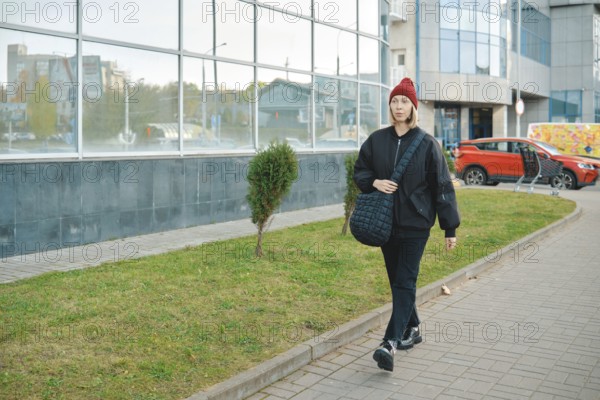A woman strolls along a wide walkway surrounded by a mix of urban buildings and greenery. She wears a black jacket, black pants, and a red beanie while carrying a shoulder bag