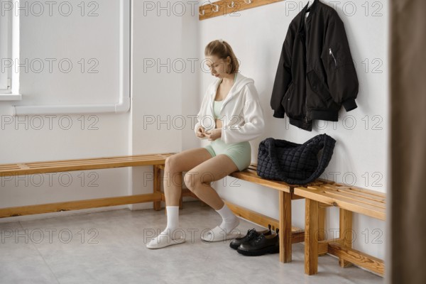 A young woman in a light green outfit sits on a wooden bench in a gym locker room. She is putting on her hoodie while a black jacket hangs nearby. A gym bag rests on the bench beside her