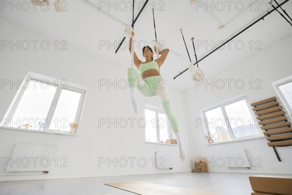 A woman in green activewear performs stretching exercises with the support of a hammock. She is in a spacious, well-lit studio with large windows, showcasing her flexibility and strength