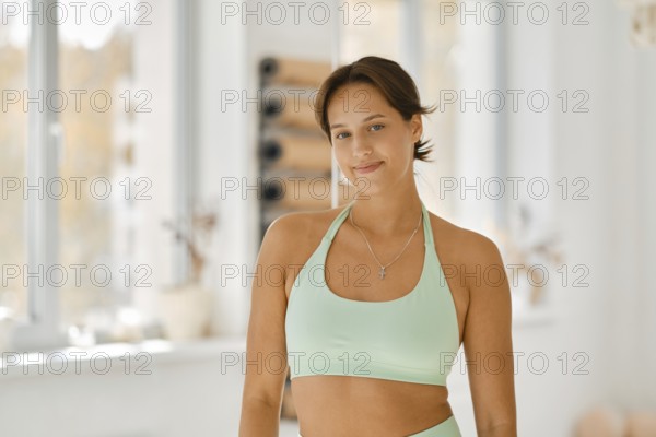A woman wears a light green workout outfit while smiling in a well-lit fitness studio. The space features large windows and fitness equipment, creating an inviting atmosphere for exercise