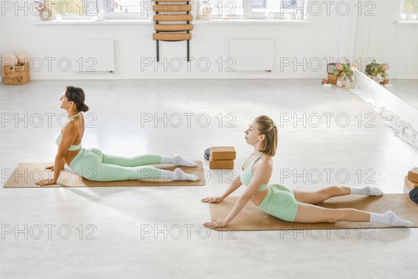 Two women are engaged in yoga practice on mats in a sunlit studio. They are positioned in deep stretches, focusing on their breathing and movements to enhance flexibility and strength
