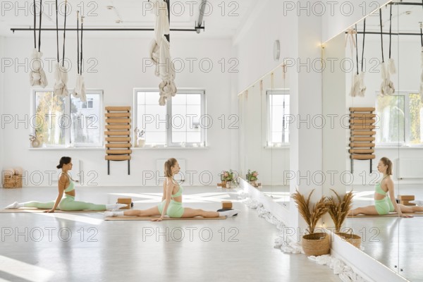 In a spacious yoga studio filled with natural light, two women perform stretches on mats. The room features large mirrors, calm decor, and hanging ropes