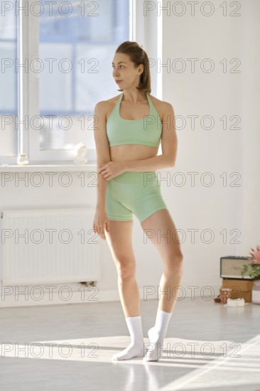 A woman in a green outfit stands gracefully in a well-lit yoga studio. She appears focused and serene, with a calm atmosphere around her. Natural light floods the space, creating a peaceful ambiance