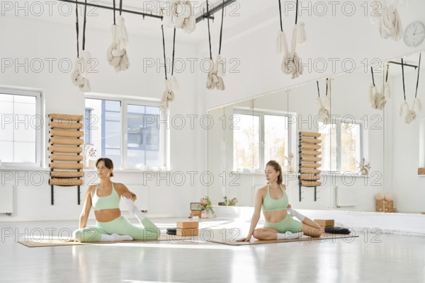 Two women are practicing stretching in a spacious and well-lit studio. They focus on different poses while enjoying the calm atmosphere created by natural light and greenery