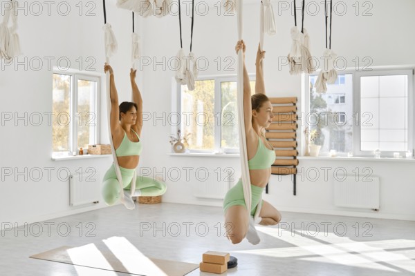 Two women practice fitness stretching with hammocks in a bright studio. They focus on flexibility and core strength while using the suspended fabric
