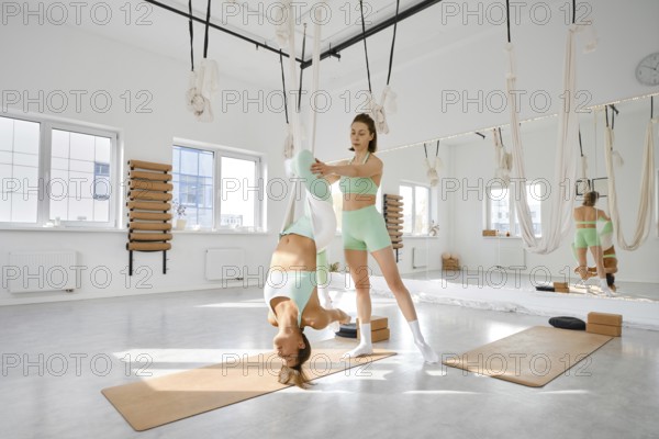 Two fitness enthusiasts are participating in a stretching session using a hammock in a bright, airy studio