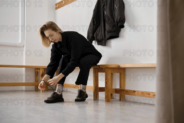 In a bright and simple locker room, a young woman sits on a wooden bench, focusing on tying their shoes. A black jacket hangs on the wall nearby, creating a casual atmosphere