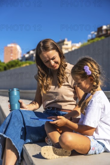 Mother and daughter smiling, sitting together on steps enjoying time outdoors, holding a reusable cup and interacting with a digital tablet, learning and connecting in sunlight