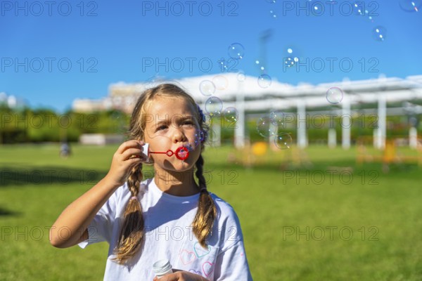 Child with pigtails enjoying summer playtime outdoors, actively creating and watching soap bubbles float in the bright blue sky above a green grass field