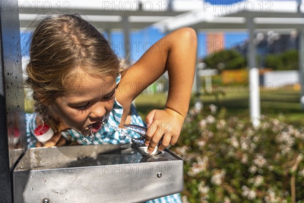 Little girl bending to drink cool water from a stainless steel park fountain on a sunny summer day, enjoying refreshment, hydration and playful outdoor fun amidst greenery