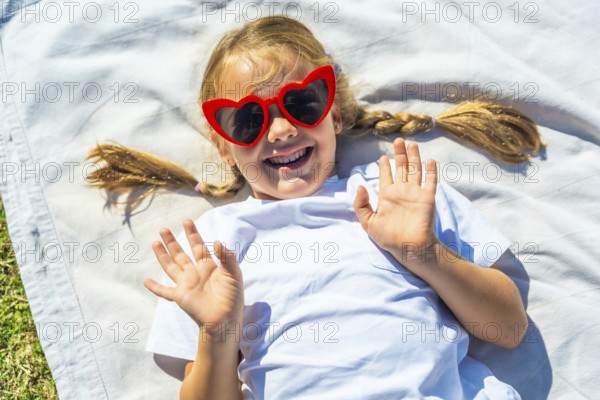 Little girl with braided blonde pigtails smiling and waving from a white blanket in the park, enjoying sunny summer fun and carefree childhood joy during a warm day