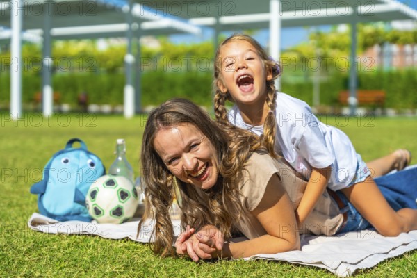 Mother and daughter bonding and having fun at a picnic in the park, the child playfully riding on her mother's back, both smiling and laughing, expressing happiness and joy