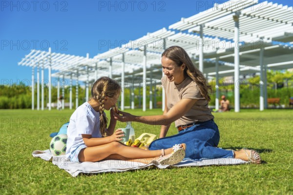 Mother and daughter enjoying a sunny summer picnic on green grass, sharing food and snacks while relaxing outdoors under a blue sky, fostering family bonding