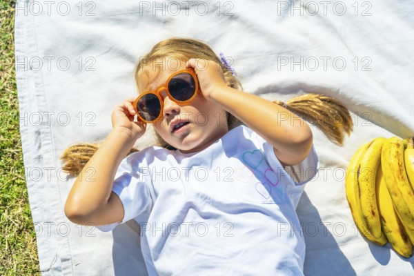 Young girl lying on a white blanket in green grass, adjusting orange sunglasses and smiling while enjoying a sunny picnic with bananas and a relaxed, carefree summer vibe