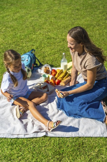 Mother and daughter share food and laughter on a picnic blanket in green grass, enjoying sunny park time, bonding over healthy snacks and carefree summer moments together