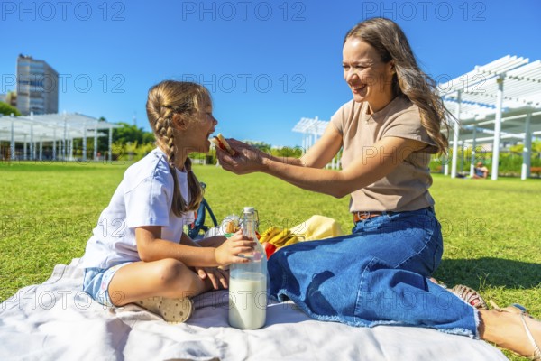 Young mother feeding her little daughter a sandwich during a sunny summer picnic on a blanket in the park, sharing tender moments of bonding, joy and outdoor relaxation