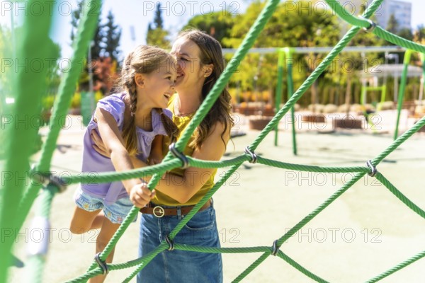 Mother supporting her happy young daughter climbing on a green rope playground structure, sharing a joyful moment of connection and fun outdoors on a sunny day