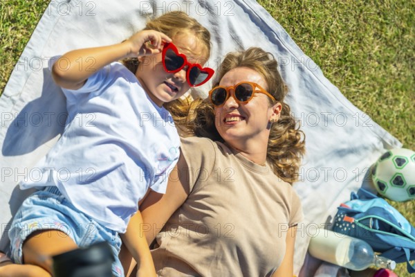 Mother and daughter lying on a picnic blanket in green grass, wearing sunglasses and smiling together outdoors on a sunny summer day, enjoying relaxed bonding and joy
