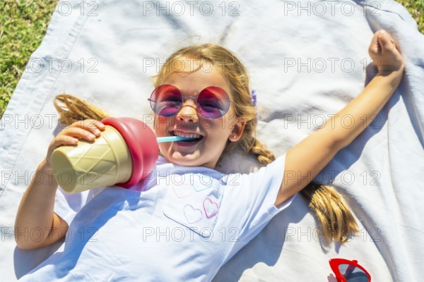 Young girl lying on a blanket in the grass on a sunny day, laughing while drinking from an ice cream shaped thermos bottle and wearing colorful sunglasses during an outdoor activity