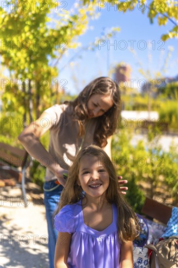 Mother is combing her daughter's long hair with a comb while the happy girl is looking at the camera and smiling, enjoying a summer day in a sunny park