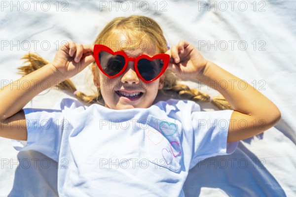 Young child lying outdoors on a white blanket, expressing joy and playing with vibrant red heart shaped sunglasses, embodying summer fun and carefree days