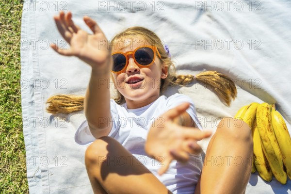 Young happy girl relaxing on a white blanket in the grass, wearing orange sunglasses and white t shirt, excitedly reaching hands towards the camera on a sunny summer day
