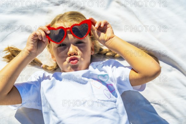 Young blonde girl lying on a white blanket making a fun kiss face while wearing red heart shaped sunglasses, expressing childhood joy and a carefree summer mood