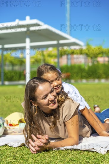 Mother and daughter lying together on a blanket, smiling and hugging, enjoying summer day outdoors in a green park, symbolizing family love, happiness, parenting, and childhood
