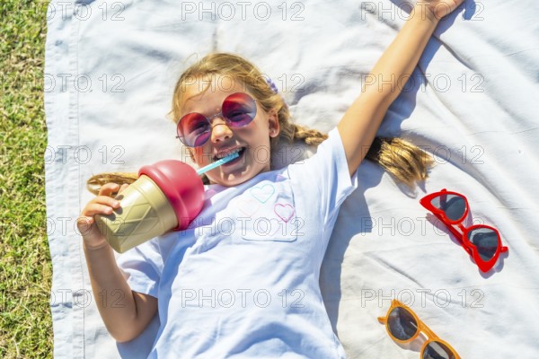 Young girl lying on a white blanket in green grass, smiling while drinking from an ice cream shaped cup through a blue straw, enjoying summer fun outdoors