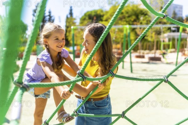 Mother and daughter bonding through playful activity, climbing together on a green rope net structure at a sunny outdoor park, enjoying happy family time