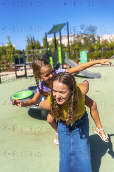 Mother gives daughter a piggyback at a sunny playground, both laughing and smiling as they enjoy playful summer fun, bonding and carefree outdoor family time