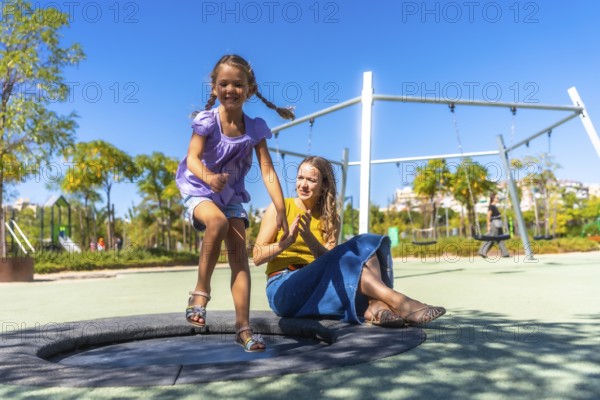 Daughter jumps on a round trampoline at a sunny park playground while mother claps nearby, sharing joyful family bonding and carefree outdoor fun on a warm day