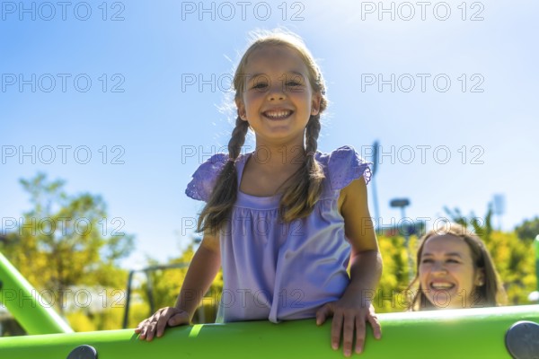 Young girl with pigtails smiling at the camera while her mother watches from playground equipment on a sunny day, enjoying outdoor family fun and carefree childhood joy
