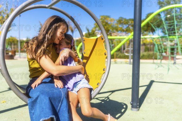 Mother and daughter share a warm hug and bright smiles on a sunny playground, enjoying playful bonding, affection and joyful family time outdoors