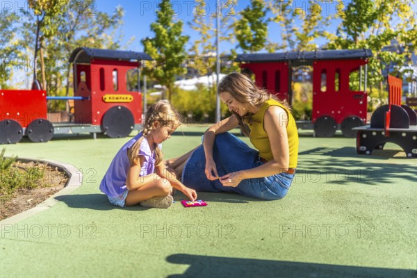 Mother and daughter bonding outdoors, sitting on the soft playground surface and concentrating on solving a small geometric puzzle together during a warm sunny day