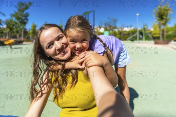 Young mother and daughter smiling at the camera while hugging on a sunny park playground, enjoying a joyful, candid moment of bonding and play outdoors during summer