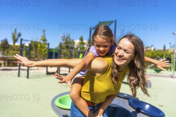 Happy mother giving her laughing daughter a piggyback ride on a sunny day in a modern public playground, symbolizing family bonding and playful childhood moments
