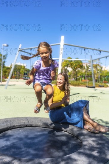 Mother and daughter experiencing a joyful moment together, with the young girl enthusiastically jumping on a playground trampoline under the bright summer sky while her mother watches happily