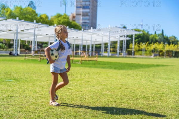 Young girl walking across a sunny green park field, carrying a small soccer ball and backpack, smiling as she enjoys a carefree summer day of play and outdoor fun