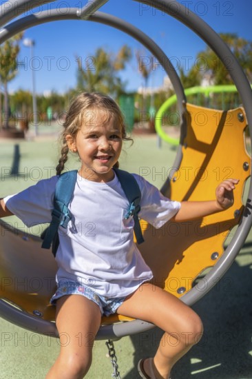 Happy little girl with a backpack smiles broadly while sitting on a swing in a vibrant outdoor playground, enjoying a sunny day of childhood and play