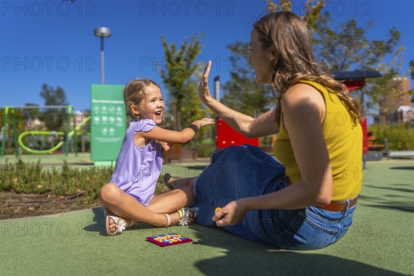 Mother and daughter sitting on playground ground, smiling and exchanging a joyful high five while playing a game on a sunny summer day