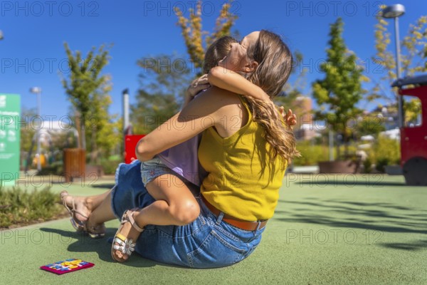Mother and daughter embracing with affection on a soft ground surface at an outdoor playground, enjoying a moment of connection and family love on a sunny day