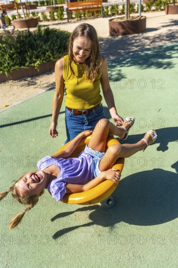 Mother standing, smiling, and gently pushing her happy daughter on a yellow spinning toy, both enjoying playtime and summer fun at a park playground on a sunny day