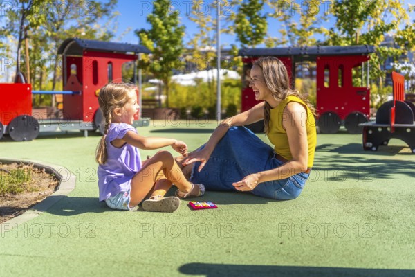 Mother and daughter sitting on soft flooring at a public playground, laughing happily during a sunny day, sharing a bonding moment while playing together